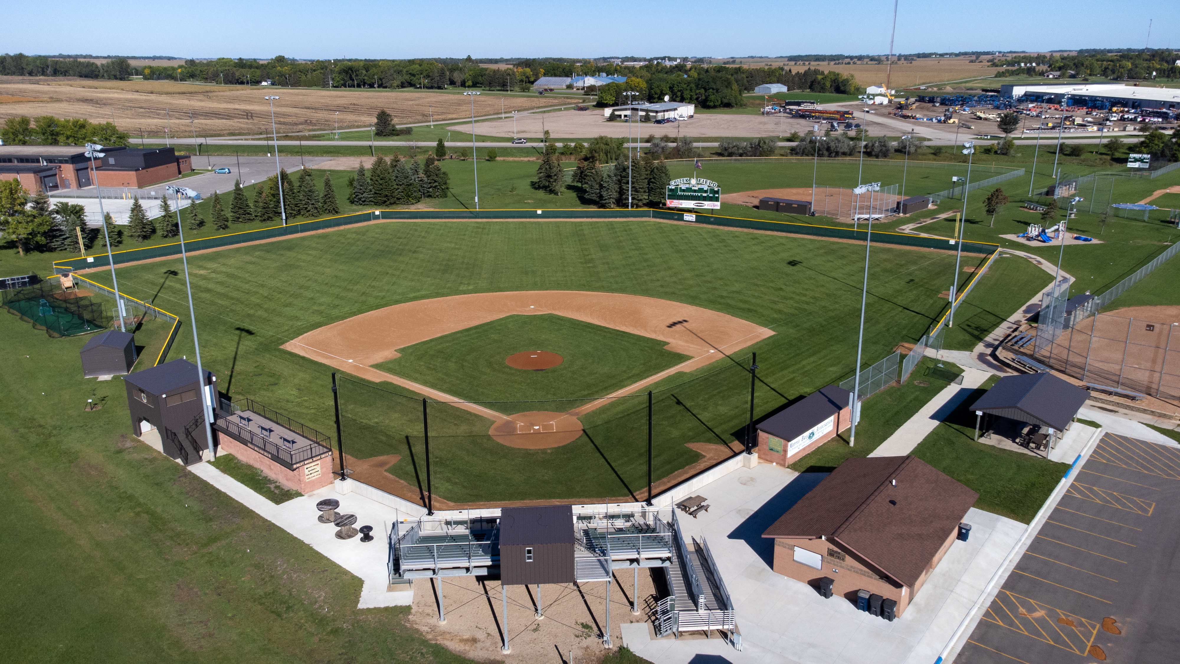 Aerial daytime view of a baseball stadium with a well-maintained diamond, bleachers, dugouts, and surrounding athletic fields set against farmland and rural landscape.